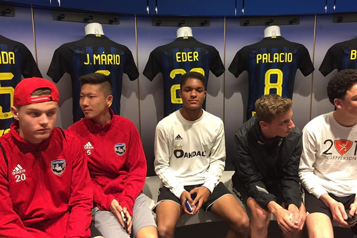 Soccer players from Marlyand sitting in a locker room in front of a row of hanging jerseys at the San Siro Stadium