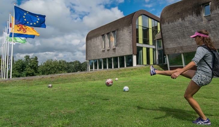 A girl kicking a soccer ball across a green lawn. Flags and buildings are surrounding the green lawn.