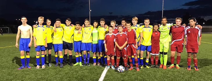 lineup of two soccer teams. One team in yellow jerseys and blue shorts, and the other team in all red jerseys, shorts and socks.