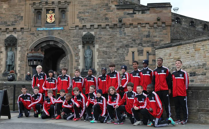 Two rows of boys posing in front of an Edinburgh Castle. The boys are wearing red and black soccer uniforms.