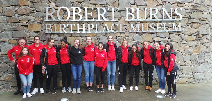 Girl soccer team standing in front of a rock wall inscribed with Robert Burns Birthplace Museum